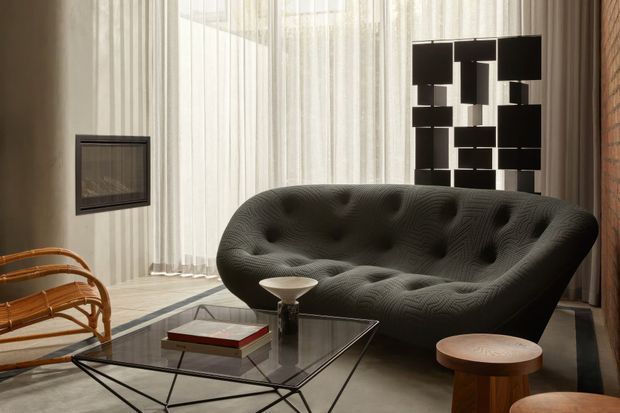 A sophisticated living room with a deeply tufted dark sofa, glass-and-metal coffee table, sculptural black wall artwork, woven lounge chair, and small wooden stools. Full-height sheer curtains diffuse daylight, while exposed brick and pale vertical wall finishes add textural contrast.