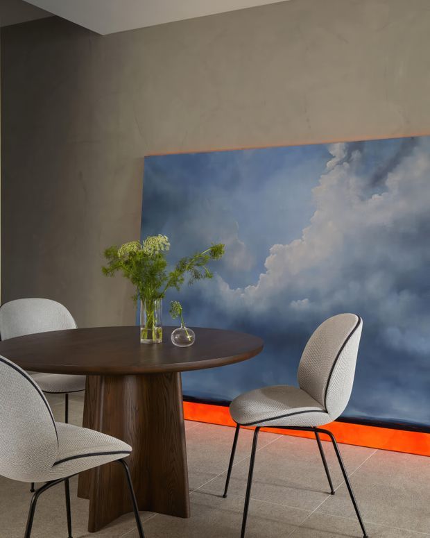 A refined dining area featuring a round dark wood pedestal table and upholstered dining chairs with slim dark metal legs. The space has smooth warm grey walls, large-format grey floor tiles, and a dramatic blue-and-white cloud artwork or wall panel with a vivid orange-red illuminated base detail.