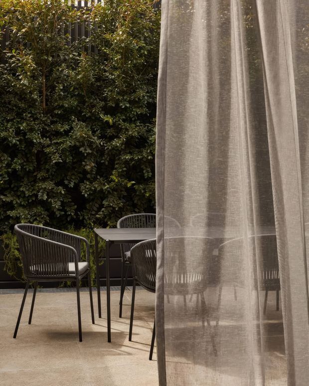 A refined outdoor terrace or patio with a dark metal dining table and woven chairs, surrounded by dense green screening plants. A sheer taupe-grey curtain hangs prominently in the foreground, partially veiling the furniture and creating layered privacy. The scene has a warm, quiet, contemporary atmosphere with stone or concrete-look paving.