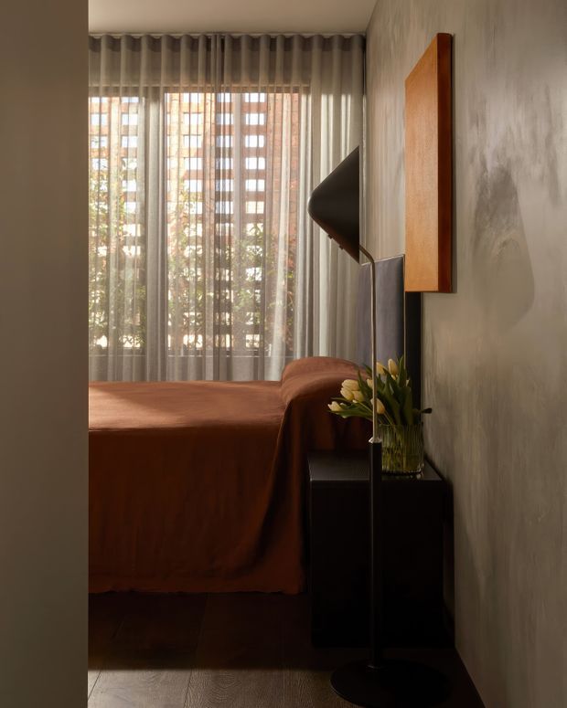 A warmly lit bedroom viewed through a doorway, featuring a low bed with rust-brown bedding, sheer floor-to-ceiling curtains, a black bedside cabinet, a sculptural black floor lamp, a vase of greenery, and a timber-framed wall artwork or mirror. The walls have a textured grey finish and the room has a refined, moody contemporary character.