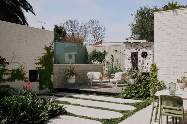 A landscaped courtyard garden with a paved outdoor entertaining area, white-painted brick walls, a small tiled raised planter, climbing greenery, and light outdoor lounge and dining furniture. Curved concrete stepping paths run through low groundcover and drought-tolerant planting, creating a serene contemporary outdoor space.