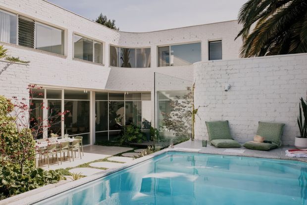 A bright white-painted brick courtyard exterior featuring a modern multi-level house with extensive glazing, a central landscaped courtyard, outdoor dining, and a swimming pool with lounge chairs. Palm planting and clean geometric lines create a relaxed resort-like setting.