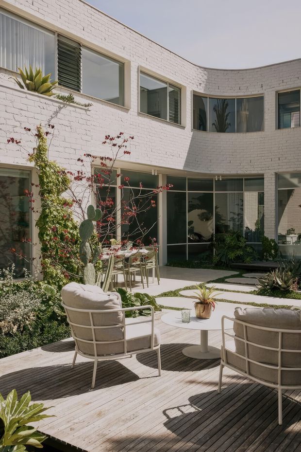 A sunlit landscaped courtyard enclosed by a curved two-storey white-painted brick building. The courtyard features a timber deck with two cushioned outdoor lounge chairs and a round table, pale paving with planted gaps, lush greenery, cacti, and glass doors and windows opening onto the surrounding rooms. The facade combines curved brickwork with extensive dark-framed glazing and exterior louvres.