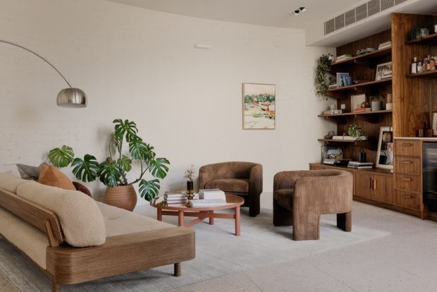 Warm, refined living room with a low-profile textured sofa, upholstered lounge chairs, a round timber coffee table, large indoor plants, and an expansive built-in timber shelving and cabinetry wall. A curved floor lamp, neutral walls, pale flooring, and curated shelving create a calm mid-century-inspired interior.