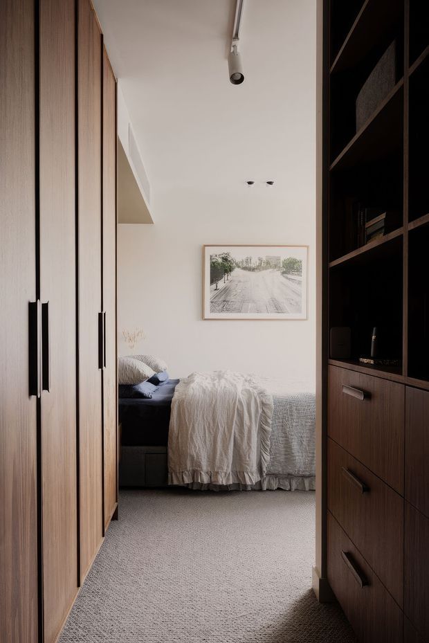 A compact, softly lit bedroom viewed through a narrow passage between built-in timber wardrobes and dark wood shelving. The room contains a bed with layered neutral bedding, a dark upholstered headboard or bench, a framed monochrome artwork, recessed and track lighting, and full-height fitted storage. The palette is warm and muted, with timber joinery, cream walls, and light grey carpeting.