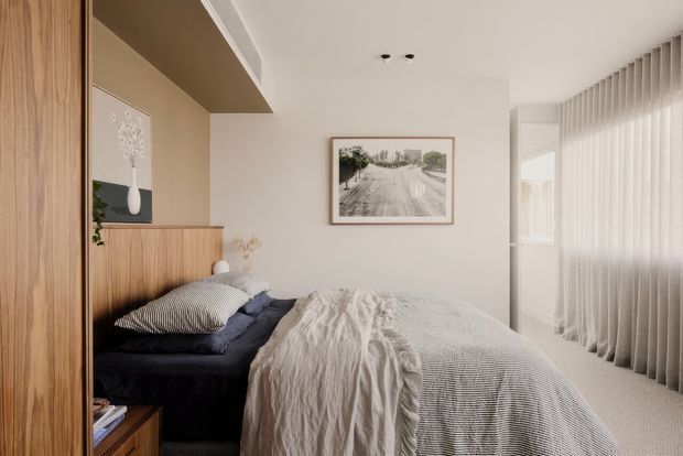 A softly lit, compact bedroom with a low dark upholstered bed, layered white and grey textured bedding, warm timber built-in cabinetry and a bedside ledge, neutral beige walls, framed artwork, and full-height sheer curtains over the window. The room has a calm, minimalist contemporary feel with warm natural materials.