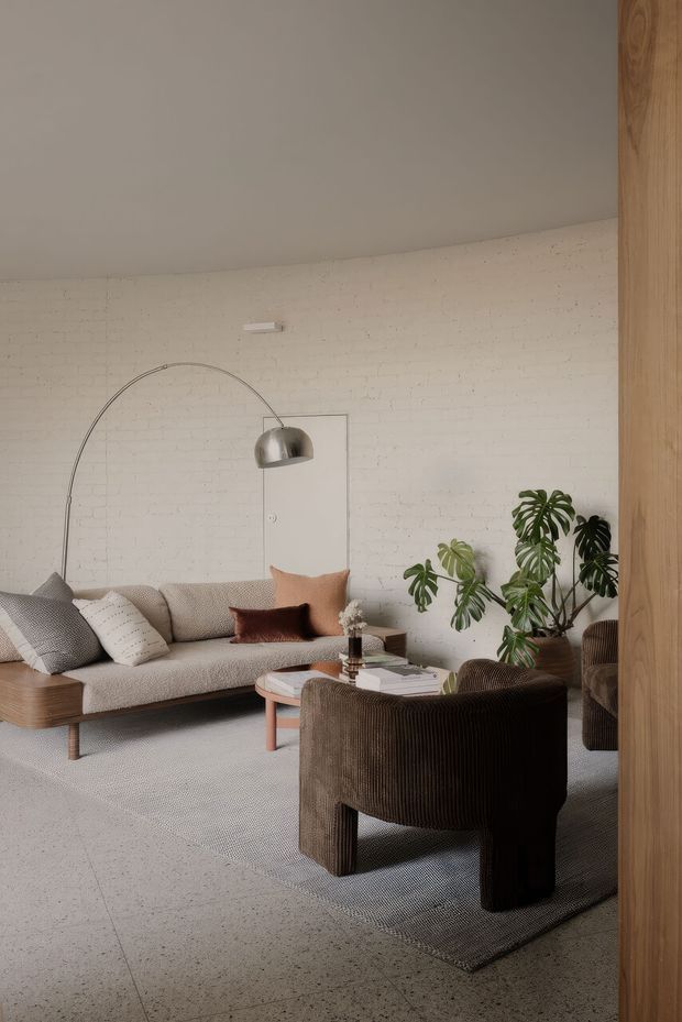 A warm, refined living room with a curved white-painted brick wall, light terrazzo flooring, a neutral textured sofa, a dark ribbed lounge chair, a low rounded coffee table, indoor tropical plants, and a large arched floor lamp. Timber wall panelling or a timber-clad opening frames the right side of the room.