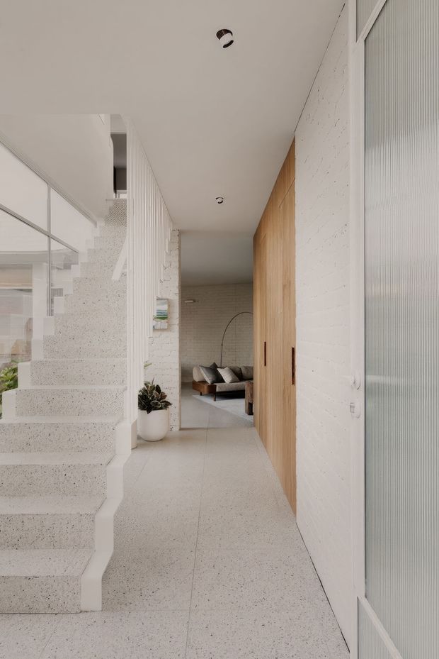 A bright minimalist residential entry hall with a terrazzo staircase and matching terrazzo floor. White painted brick walls, vertical white balustrade slats, pale timber built-in doors, and a ribbed translucent glass door create a clean contemporary interior. A living room with a sofa is visible at the end of the corridor.