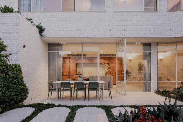 A contemporary courtyard viewed from outdoors, featuring a white-painted brick two-storey facade, large aluminium-framed glazed doors and windows, and an outdoor dining setting on a paved terrace. The interior visible through the glazing includes warm timber joinery and neutral finishes. Curved stepping stones, low groundcover, and planted borders soften the courtyard landscape.