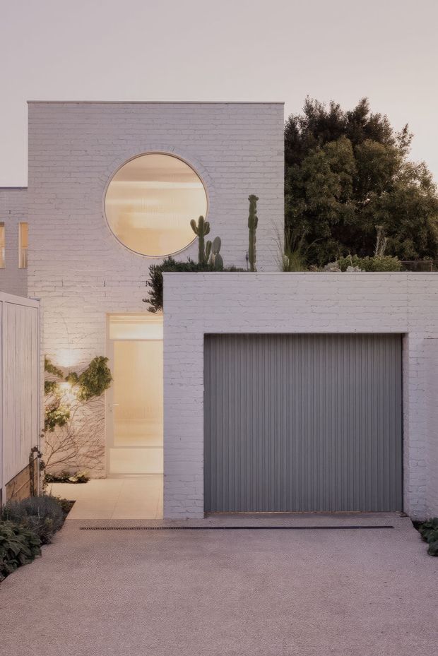 Exterior view of a refined contemporary residence with pale painted brick walls, a large circular window, warm illuminated entry glazing, a ribbed dark garage door, and integrated desert-style planting with cacti.
