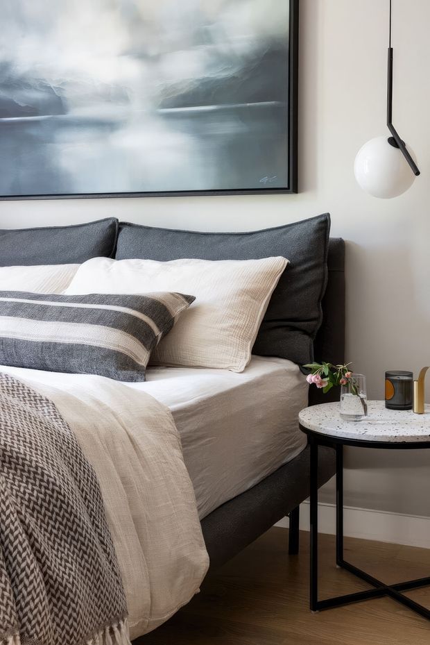 A close-up view of a neatly styled bedroom corner featuring a dark upholstered bed with layered white, cream and charcoal bedding, a large abstract artwork, and a black pendant wall light. A small round side table with a light terrazzo-like top, black metal base, candle and small vase sits beside the bed. Warm timber flooring and neutral walls create a refined, calm atmosphere.