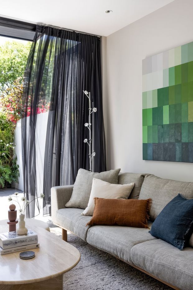 Bright contemporary living room with a large grey sectional sofa, layered neutral and rust cushions, a rounded pale wood coffee table, and a tall abstract green geometric artwork. Full-height glazing is dressed with sheer black curtains, opening to lush greenery outside. A sculptural multi-arm floor lamp and soft textured rug add detail to the refined, light-filled space.