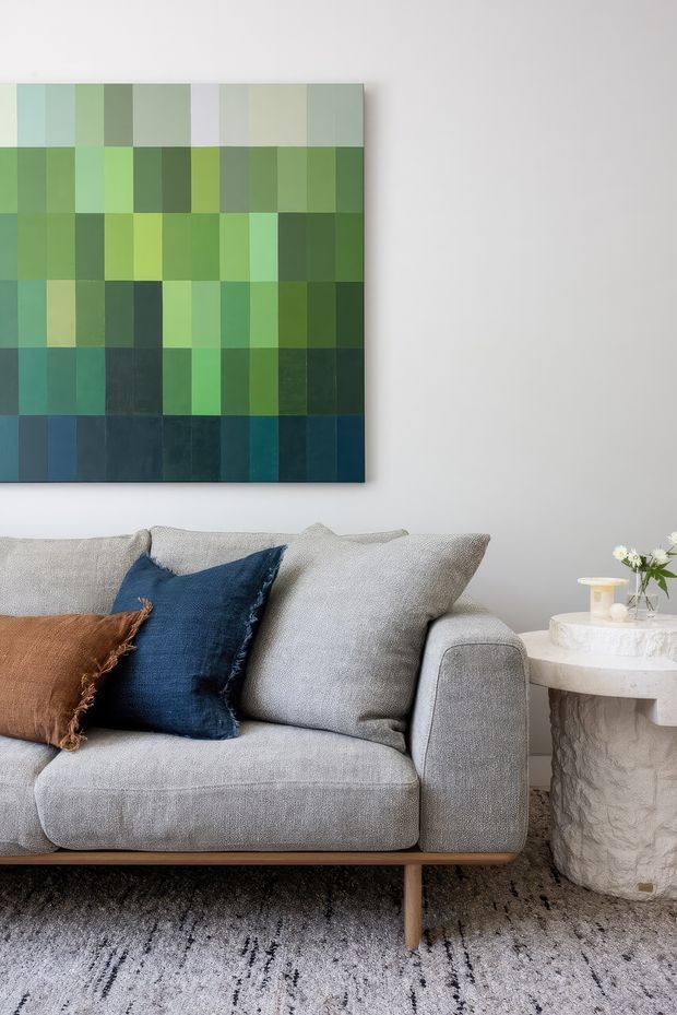A close-up of a contemporary living room featuring a light grey upholstered sofa with textured grey, navy, and rust cushions. A large geometric green and blue artwork hangs on the pale wall above the sofa. A sculptural white stone side table holds a small vase of flowers and a candle, while a neutral patterned rug covers the floor.