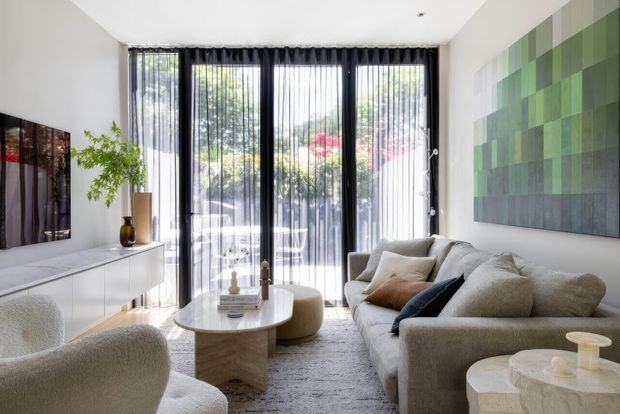 Bright contemporary living room with a neutral upholstered sectional sofa and armchair, a round stone or concrete coffee table, white built-in media cabinetry, tall black-framed glazed doors with sheer curtains, and a large green-toned abstract wall artwork. Indoor planting and garden views add a natural backdrop.