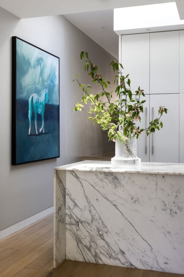 Bright contemporary interior featuring a sculptural marble-clad island or console with prominent grey veining, white built-in cabinetry, pale timber flooring, and a large blue-toned artwork. A leafy branch arrangement in a textured glass vase adds an organic accent, while a ceiling skylight or light well provides strong natural illumination.