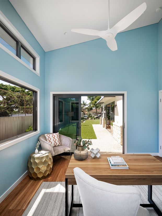 Bright contemporary home office or study with vivid pale-blue walls, a white three-blade ceiling fan, large windows, and a glazed door opening to a landscaped courtyard. A warm timber desk, white upholstered chair, patterned accent chair, metallic side table, rug, and small decorative objects create a fresh, airy workspace.