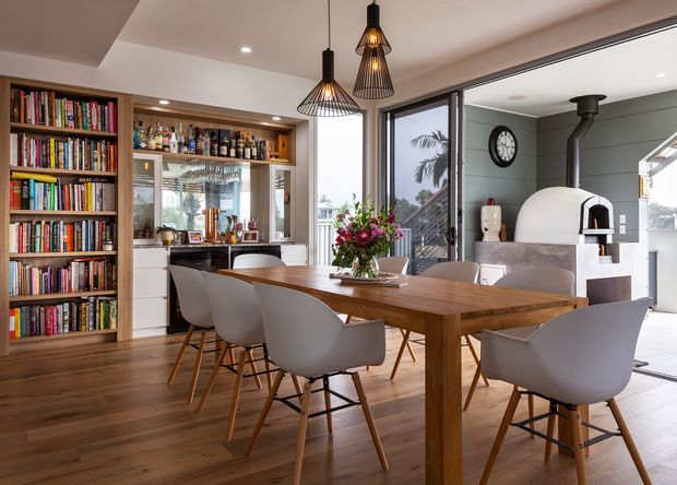 A bright open-plan dining area with a large solid timber dining table and contemporary upholstered chairs. Built-in timber bookcases and a compact bar or drinks display line the left wall, while full-height glazing opens toward an outdoor area. A sculptural white masonry pizza oven with a dark flue is visible on the right, complemented by three black geometric pendant lights and warm timber flooring.