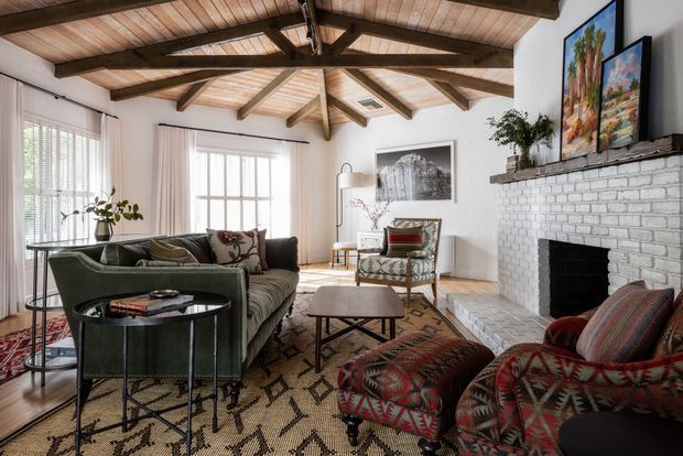A warm, eclectic living room with a vaulted timber ceiling and exposed dark-stained rafters. The room features white walls, a white-painted brick fireplace, large windows with blinds and sheer curtains, a dark green sofa, patterned armchairs and ottoman, a low geometric coffee table, and a richly patterned area rug. Framed artwork and plants add colour and character.