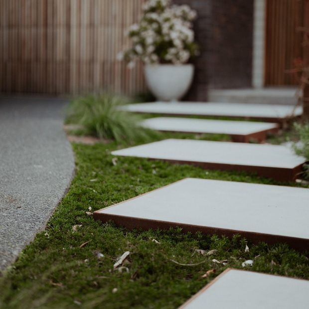 A landscaped exterior walkway with large rectangular stepping slabs set through low mossy groundcover. The pale concrete or stone pavers lead toward a timber-clad entry area, with ornamental planting and a white planter in the background.