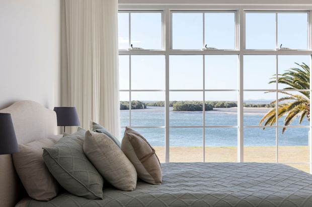 A bright bedroom with a large grid of white-framed windows overlooking a calm waterfront and sandy shoreline. The room features a neatly made bed with a grey quilted coverlet and layered neutral pillows, flanked by dark bedside lamps, with full-height curtains framing the view.