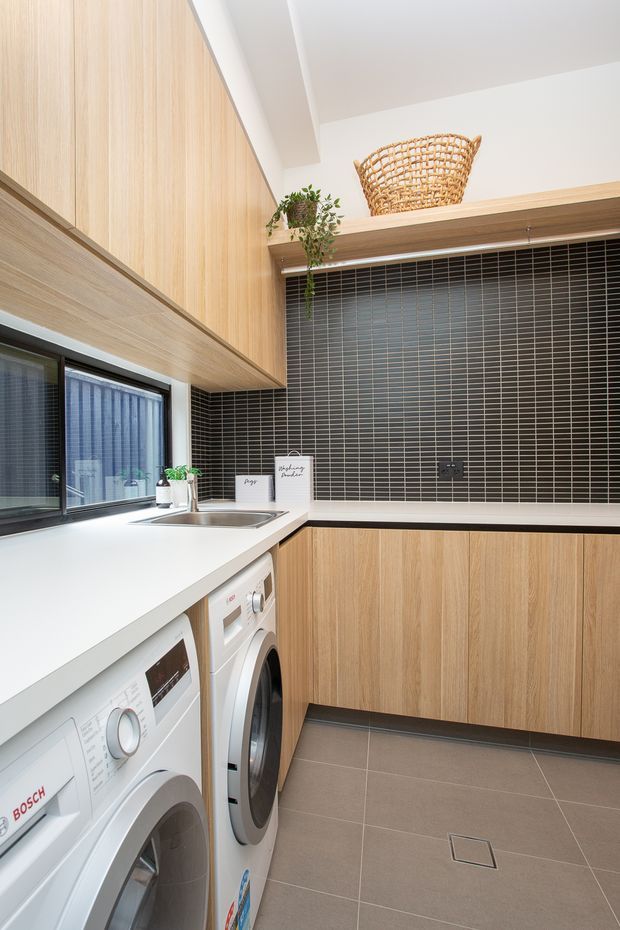 Bright, compact contemporary laundry room with stacked? side-by-side front-loading washing machines beneath a white benchtop, an inset stainless-steel sink, light timber cabinetry, and a dark charcoal small-format tiled splashback. A black-framed window provides natural light, while open shelving displays a woven basket and trailing plant. Large grey floor tiles and clean-lined cabinetry create a practical, refined fit-out.