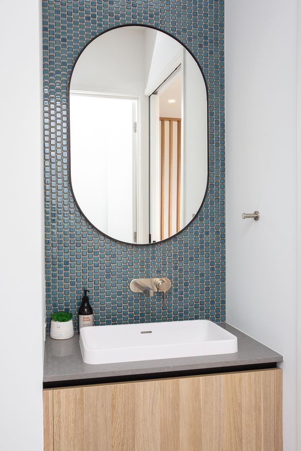 A compact contemporary bathroom vanity with a white vessel basin set on a grey benchtop above light timber cabinetry. The wall behind the basin is clad in small blue mosaic tiles and features a large rounded oval mirror with a dark frame. Brushed metal wall-mounted tapware and a simple towel hook complete the clean, bright composition.