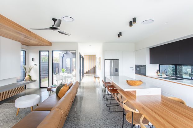 Bright open-plan contemporary interior combining a kitchen, dining area and living room. The space features a long timber dining table with stools, a white kitchen island and cabinetry, dark overhead joinery, polished terrazzo flooring, a tan leather sectional sofa, timber ceiling detailing, large glazed doors and windows, and integrated modern lighting and ceiling fans.