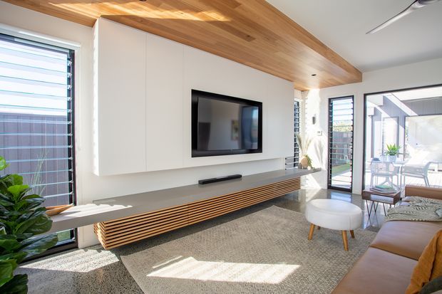 Bright contemporary living room with a wall-mounted television set into a white built-in media wall, a long floating timber-slat console, and a warm timber-lined ceiling bulkhead. Large glazed doors and windows open to an outdoor dining area, while a sectional sofa, round ottoman, rug, plants, and compact furnishings create a relaxed, sunlit setting.