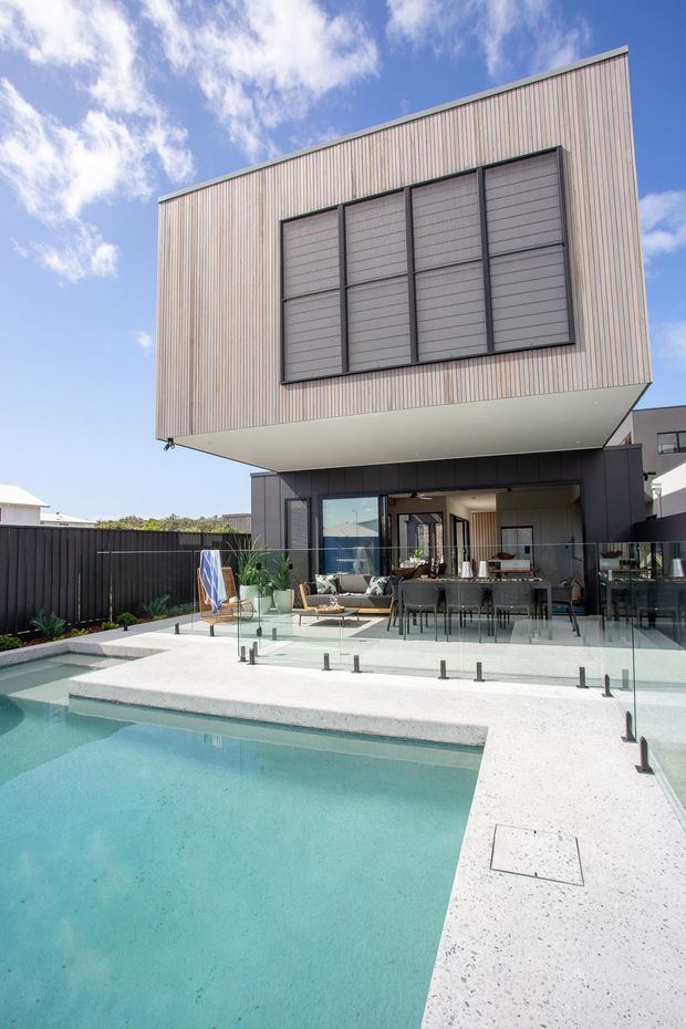 Contemporary multi-level residence viewed from the rear, featuring a dramatic cantilevered upper volume clad in light vertical corrugated panels with large dark-framed window shutters. The ground level opens onto a glazed outdoor dining and lounge terrace beside a rectangular swimming pool, with pale aggregate paving and dark boundary fencing. Bright sunny exterior with blue sky.