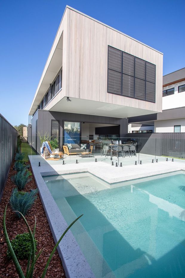 Exterior view of a contemporary multi-storey residence with a projecting upper volume clad in pale vertical timber battens. The ground level opens onto a covered outdoor entertaining area with dining furniture, seating, glazing and a balcony-like terrace overlooking a geometric swimming pool. Dark fencing, landscaped planting and neighbouring modern homes frame the scene under a clear sky.