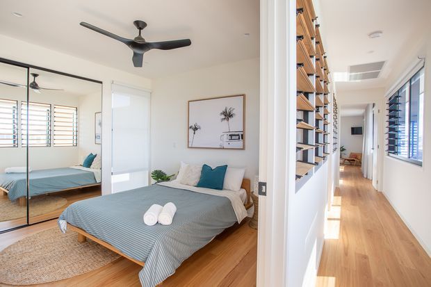 Bright contemporary bedroom viewed from an adjoining hallway, with a timber floor, neutral walls, a low-profile bed dressed in blue-grey bedding, mirrored wardrobe doors, ceiling fan, and framed artwork. The adjacent corridor features warm timber flooring, extensive built-in open shelving, louvred windows, and abundant natural light.