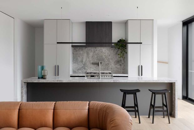 Bright contemporary open-plan kitchen viewed from the living area, featuring a large stone island with two black stools, white full-height cabinetry, dark lower cabinetry, a marble-look splashback, integrated cooktop and sink, and a partial view of a tan leather sofa. Black-framed glazing is visible at right.