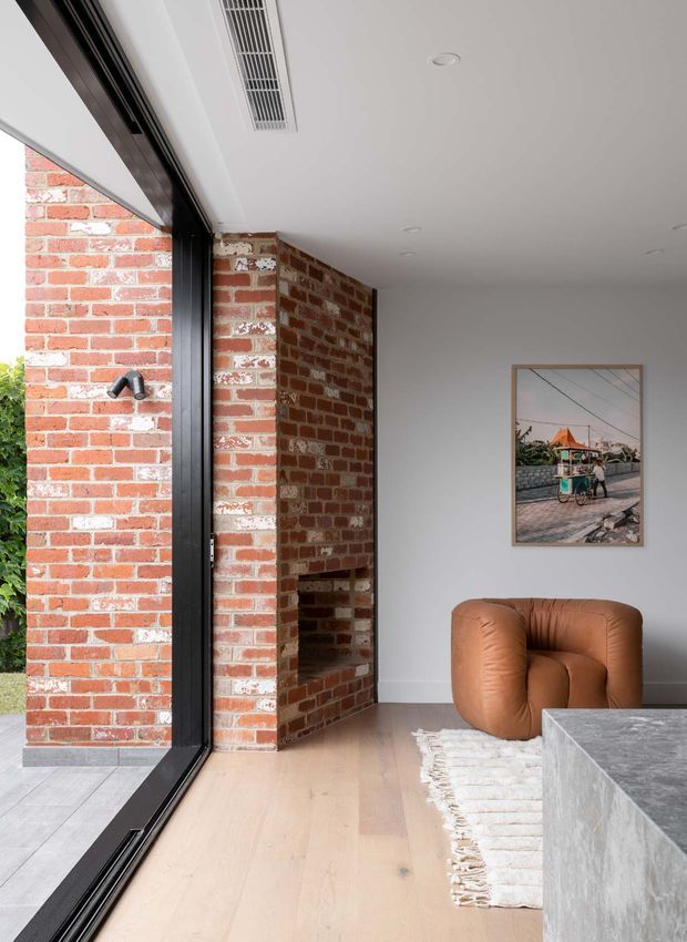 A contemporary living room with exposed red brick walls and a built-in brick fireplace, pale timber flooring, white ceiling, expansive black-framed glazing, and a sculptural tan leather armchair. A framed artwork, textured rug, and grey stone-clad coffee table add restrained warmth and contrast.