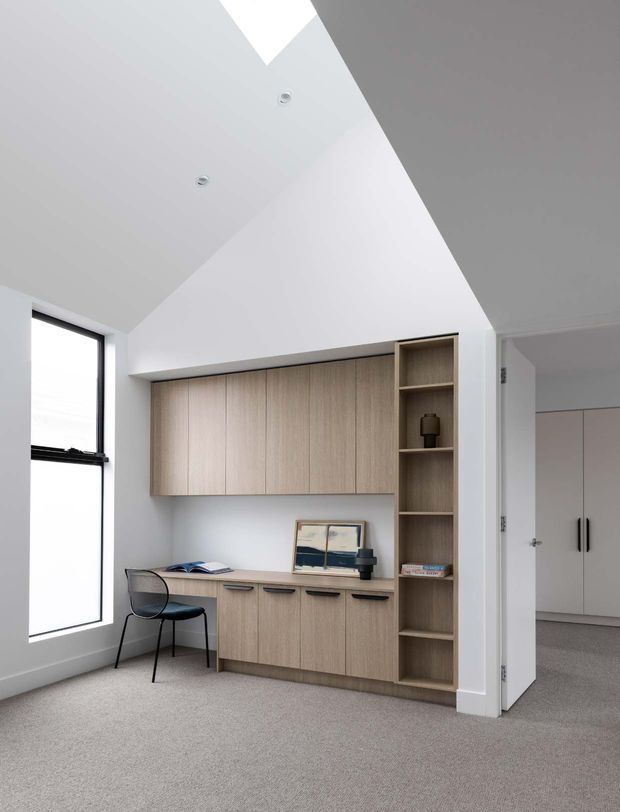 Bright minimalist home office with custom light timber built-in cabinetry, integrated desk, open shelving, and a black-framed window. The room has white walls and a sharply angled ceiling with recessed downlights, while a carpeted floor and adjoining built-in wardrobe are visible through the doorway.