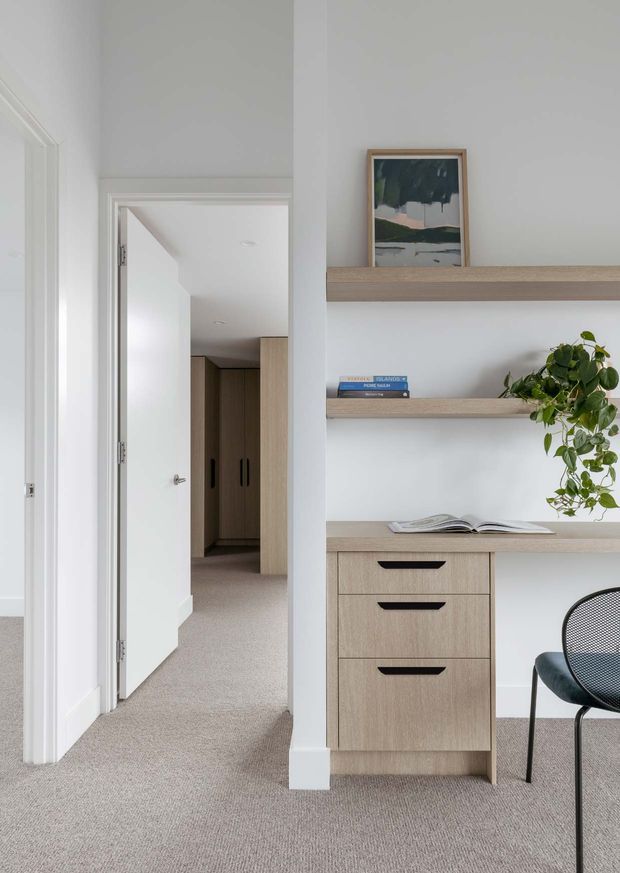 Bright, minimalist home office nook integrated into a hallway, with pale oak built-in shelving and drawers, a white desk surface, open shelving, framed artwork, books, and a trailing plant. A black mesh chair sits beside the joinery, while an open doorway leads to a carpeted hall and timber-toned cabinetry beyond.