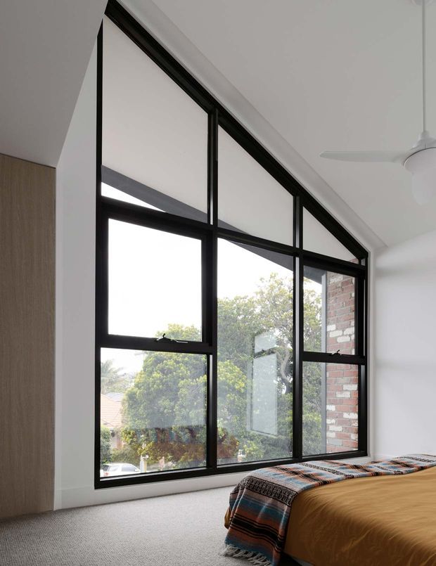 Bright contemporary bedroom with a large black-framed multi-panel window wall, white walls and ceiling, soft grey carpet, and a bed dressed in warm ochre bedding with a patterned throw. The glazing overlooks leafy suburban surroundings and reveals exposed brickwork outside.