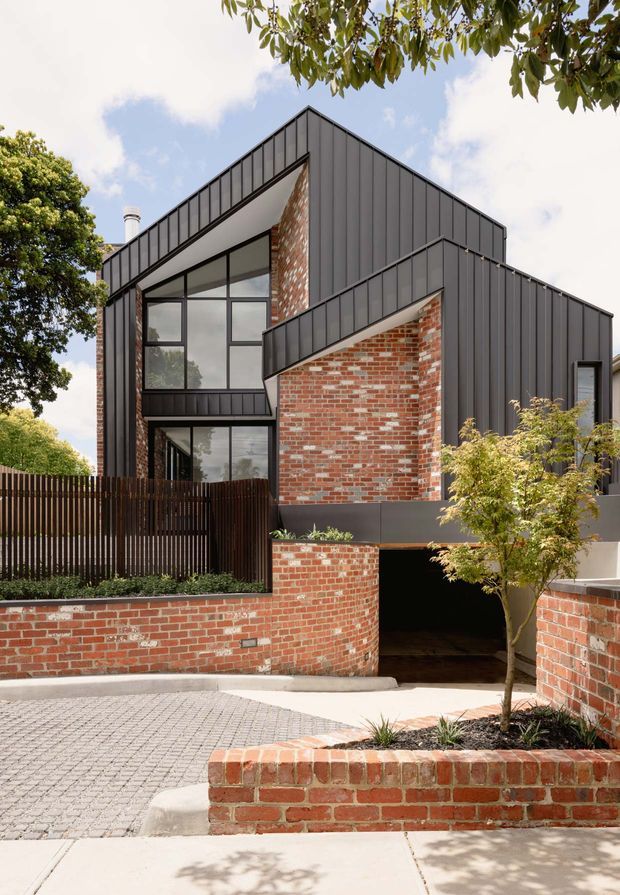 Street-level exterior view of a contemporary multi-level house with a strongly geometric gabled form. The facade combines dark vertical metal cladding, large black-framed glazed openings, and exposed red brick walls. A brick retaining wall, paved driveway, landscaping, and an open garage entrance are visible in the foreground.