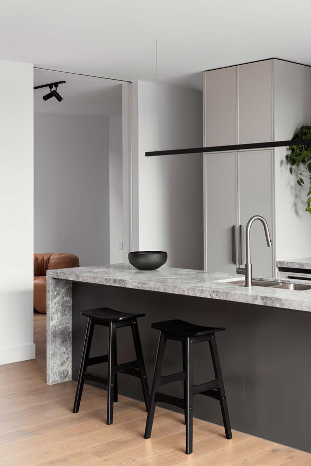 A contemporary minimalist kitchen with a large marble-look island featuring a waterfall edge, dark charcoal cabinetry, integrated sink and curved stainless-steel tap. Two black stools sit at the island beneath a slim black linear pendant light. Tall pale cabinetry provides storage, while light timber flooring and white walls create a clean, restrained interior. A glimpse of a living area is visible through the opening.