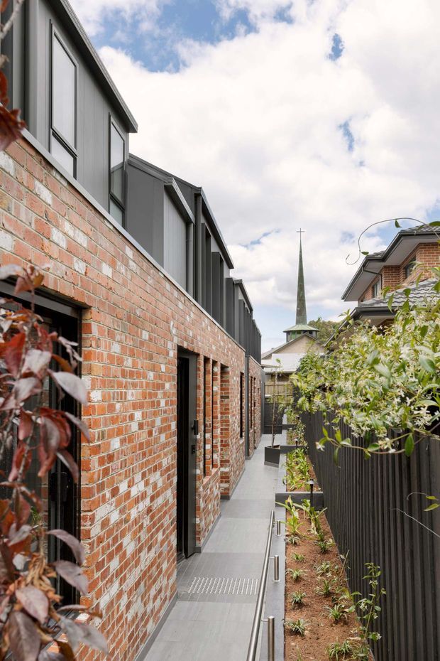 A narrow landscaped external walkway runs beside a long red-brick building with dark grey metal-clad upper levels and black-framed doors. A black vertical-slat fence and planted garden border the opposite side, while a church spire is visible in the background beneath a partly cloudy sky.