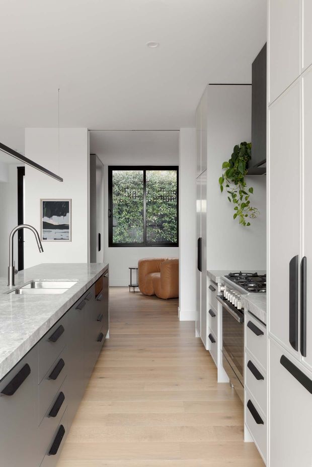 A narrow, contemporary galley kitchen viewed toward an adjoining living area. The kitchen has dark grey lower cabinetry and tall white cabinetry with long black handles, a pale stone benchtop, integrated stainless-steel appliances, a gas cooktop, undermount sink, timber flooring, and a black-framed window overlooking greenery. A tan leather sofa and trailing plant are visible beyond the kitchen.