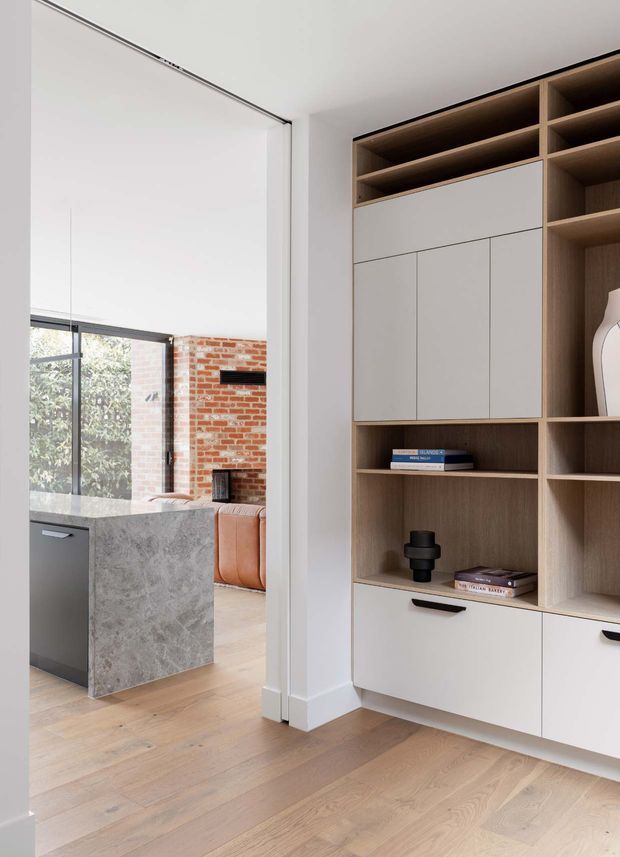 Bright contemporary interior showing a full-height built-in timber and white cabinetry wall with open shelving, enclosed cupboards and drawers. A large opening reveals part of an adjacent kitchen and living area, including a grey stone island, black-framed glazing, exposed red brickwork and a tan leather sofa. Light timber flooring and white walls create a clean, warm finish.