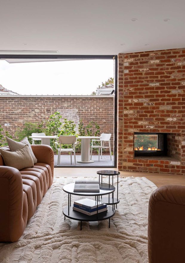 A warm contemporary living room with exposed red brick walls, a built-in fireplace, curved tan leather seating, layered round black coffee tables, and a textured cream rug. Large sliding or folding glazing opens the room to a small paved courtyard with an outdoor dining setting.