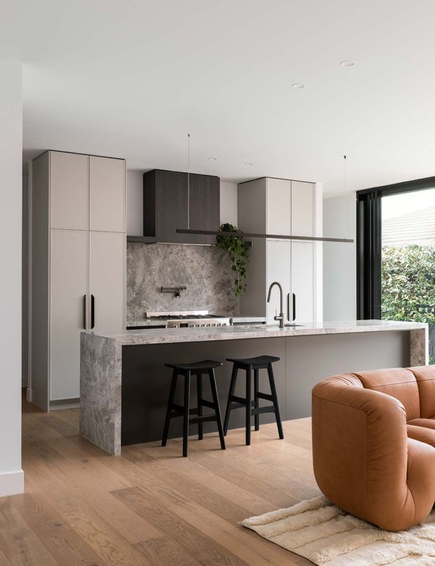 A refined contemporary open-plan kitchen with a large stone island and integrated seating, tall pale cabinetry, a dark timber-toned rangehood cabinet, and a textured stone splashback. Light timber flooring and a caramel leather sofa connect the kitchen to the adjacent living area. Black stools, tapware, window frames, and a slim linear pendant provide contrast, while a large glazed opening brings in daylight and greenery.