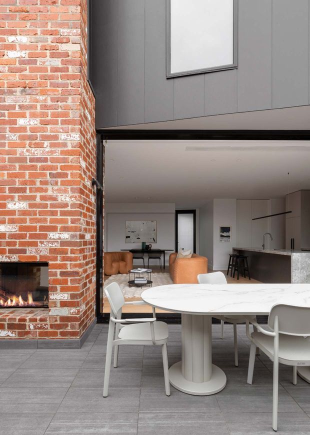 A contemporary open-plan residential interior viewed from a covered outdoor or threshold area. A substantial exposed red brick fireplace with a lit fire anchors the left side, while a large white oval dining table with sculptural pedestal and white chairs occupies the foreground. Behind it is a minimalist living area with caramel lounge chairs and a compact coffee table, opening into a sleek kitchen with a dark stone island, integrated cabinetry and bar stools. Grey tiled flooring, white walls and dark grey vertical exterior cladding around the opening create a refined, warm-modern aesthetic.