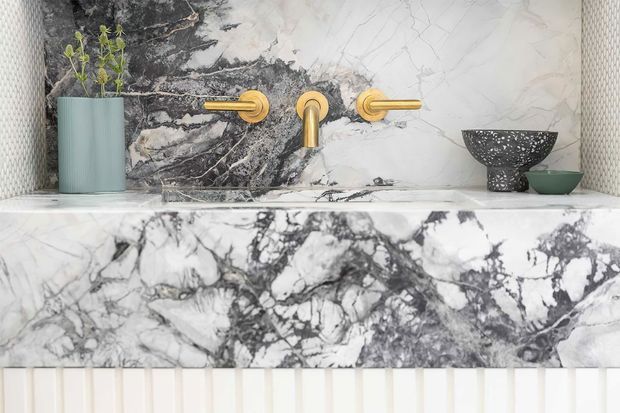 Close-up of a refined bathroom vanity or basin installation featuring dramatic white-and-grey veined marble across the wall backsplash and thick countertop. Brushed gold wall-mounted tapware provides a warm accent, with a pale green vessel, patterned dark bowl, and textured wall finish adding decorative detail. Vertical ribbed detailing is visible below the counter.