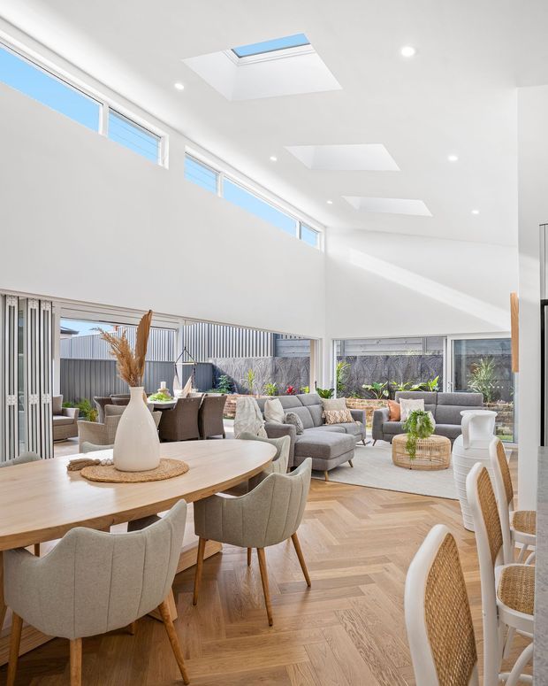 Bright open-plan living and dining area with a double-height ceiling, extensive glazing, multiple skylights, and light herringbone timber flooring. A large timber dining table with upholstered chairs occupies the foreground, while a grey sectional seating area and indoor plants sit beyond. The interior has a clean, contemporary feel with abundant natural light and neutral furnishings.