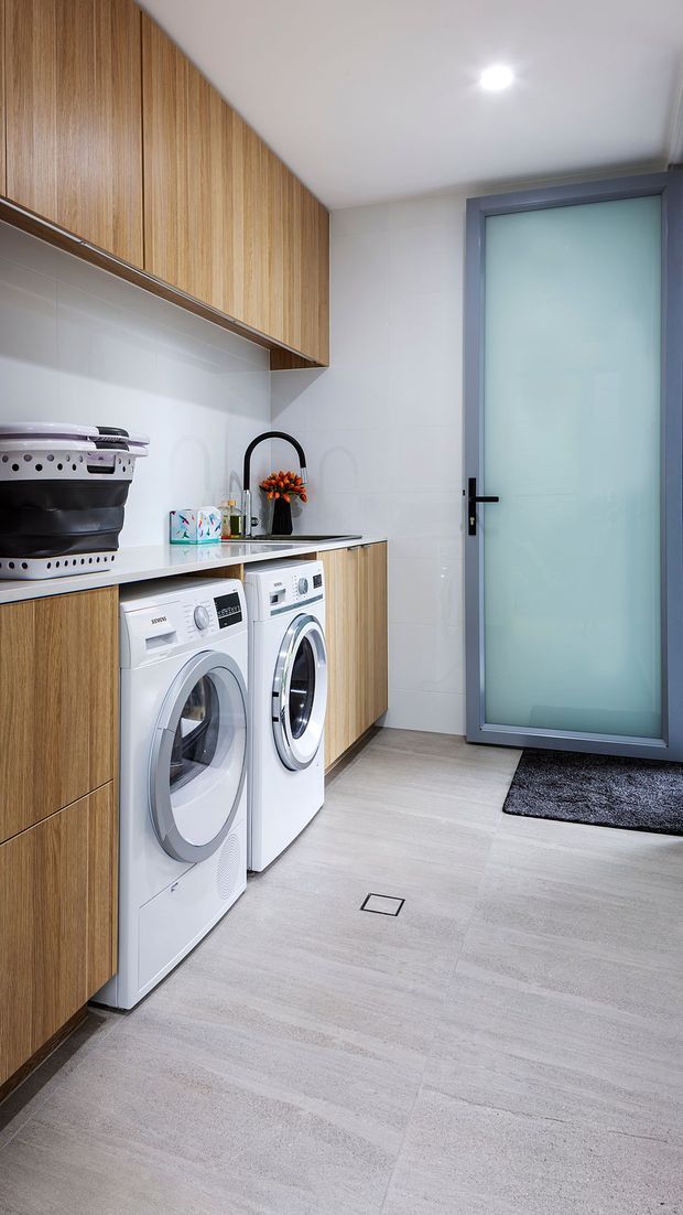 A compact, clean laundry room with floor-to-ceiling light timber cabinetry, white tiled walls, a pale grey tiled floor, white front-loading washer and dryer, and a dark benchtop with integrated sink. A frosted-glass door with a grey frame provides access, while black tapware and hardware add contrast.