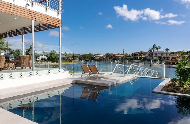 Bright waterfront residential exterior featuring a modern multi-level home with expansive glazing, horizontal timber screening, balconies, and a tiled pool terrace. A dark blue swimming pool sits beside a canal or lagoon, with lounge chairs, glass balustrades, and a small bridge-like pool edge. The scene has a relaxed tropical coastal atmosphere under a clear blue sky.