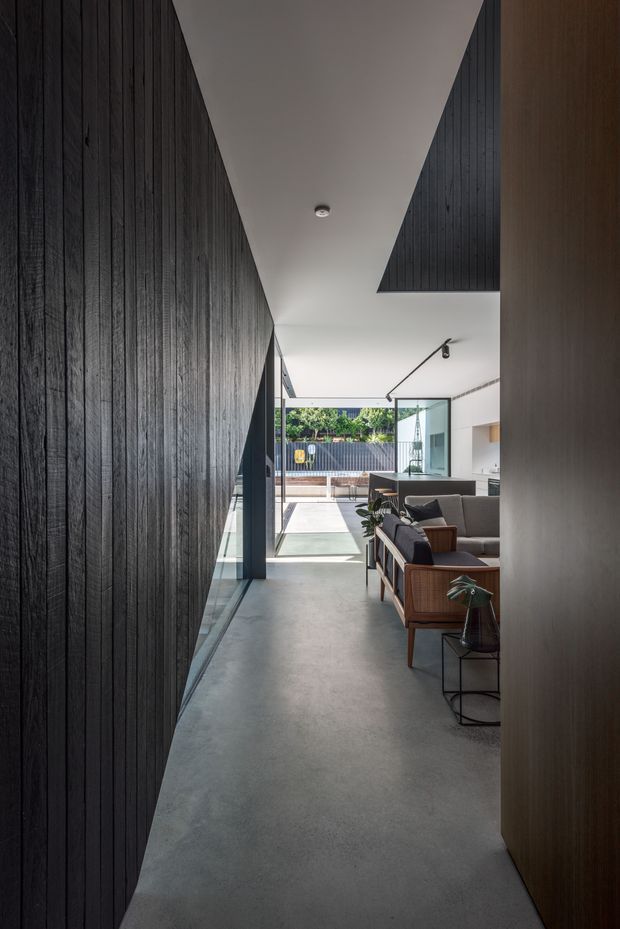 A narrow modern interior passage opens into a bright living area and courtyard. The space features a dark vertically timber-lined wall, smooth grey concrete flooring, white ceilings, timber joinery, large glazed openings, and mid-century-inspired lounge chairs. A partially visible outdoor terrace and planted garden are seen beyond the glazing.