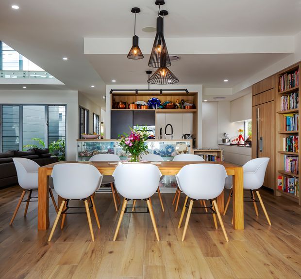 Bright open-plan dining and kitchen interior with a large light timber dining table surrounded by white moulded chairs, pendant lights above, timber flooring, extensive built-in shelving, and a kitchen with island cabinetry in the background. A living area and stair void are visible to the left.